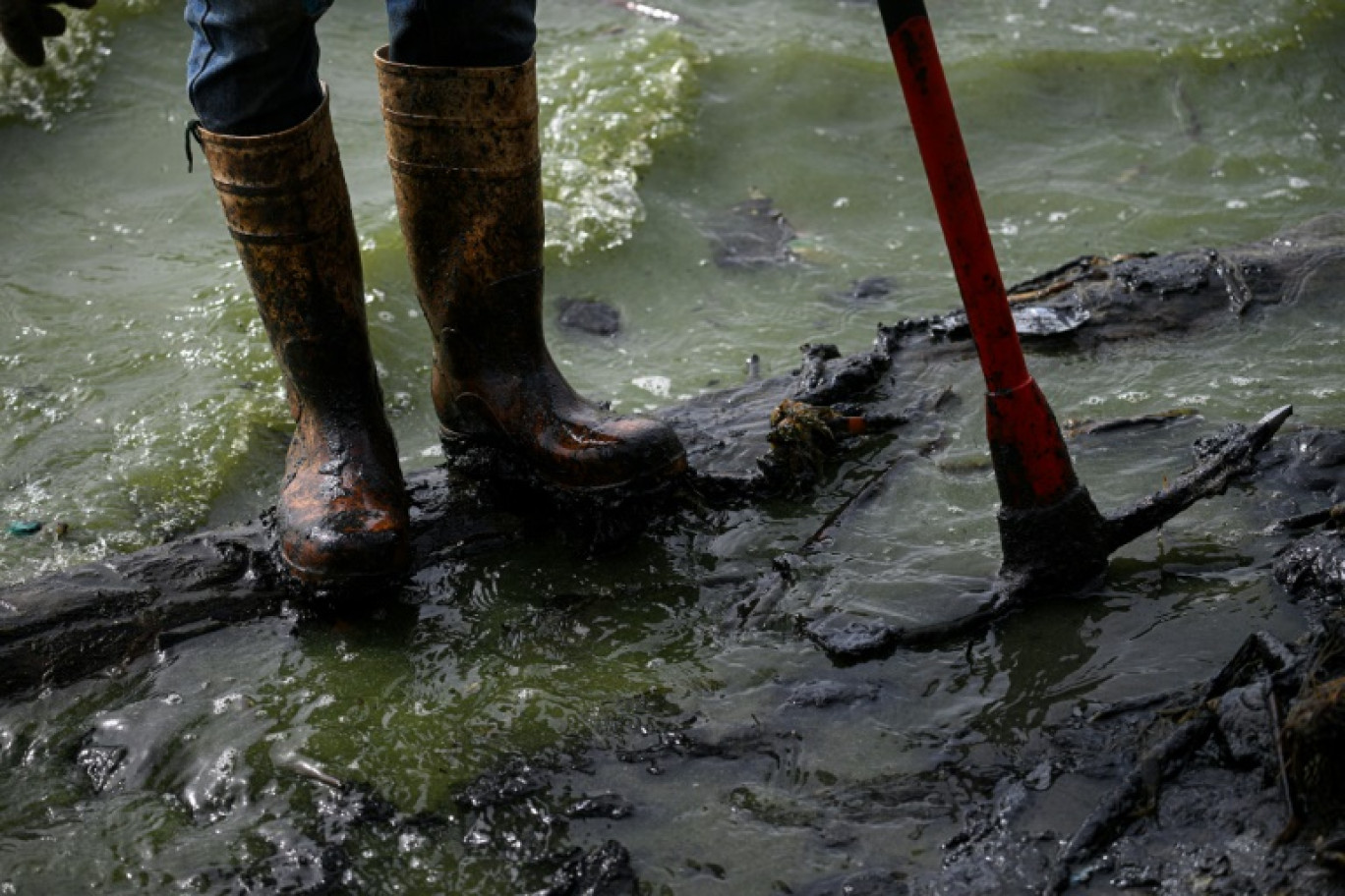 Un pêcheur aux bottes couvertes de pétrole se tient sur une rive polluée du lac Maracaibo, dans l'État de Zulia, au Venezuela, le 11 juillet 2024 © Federico PARRA