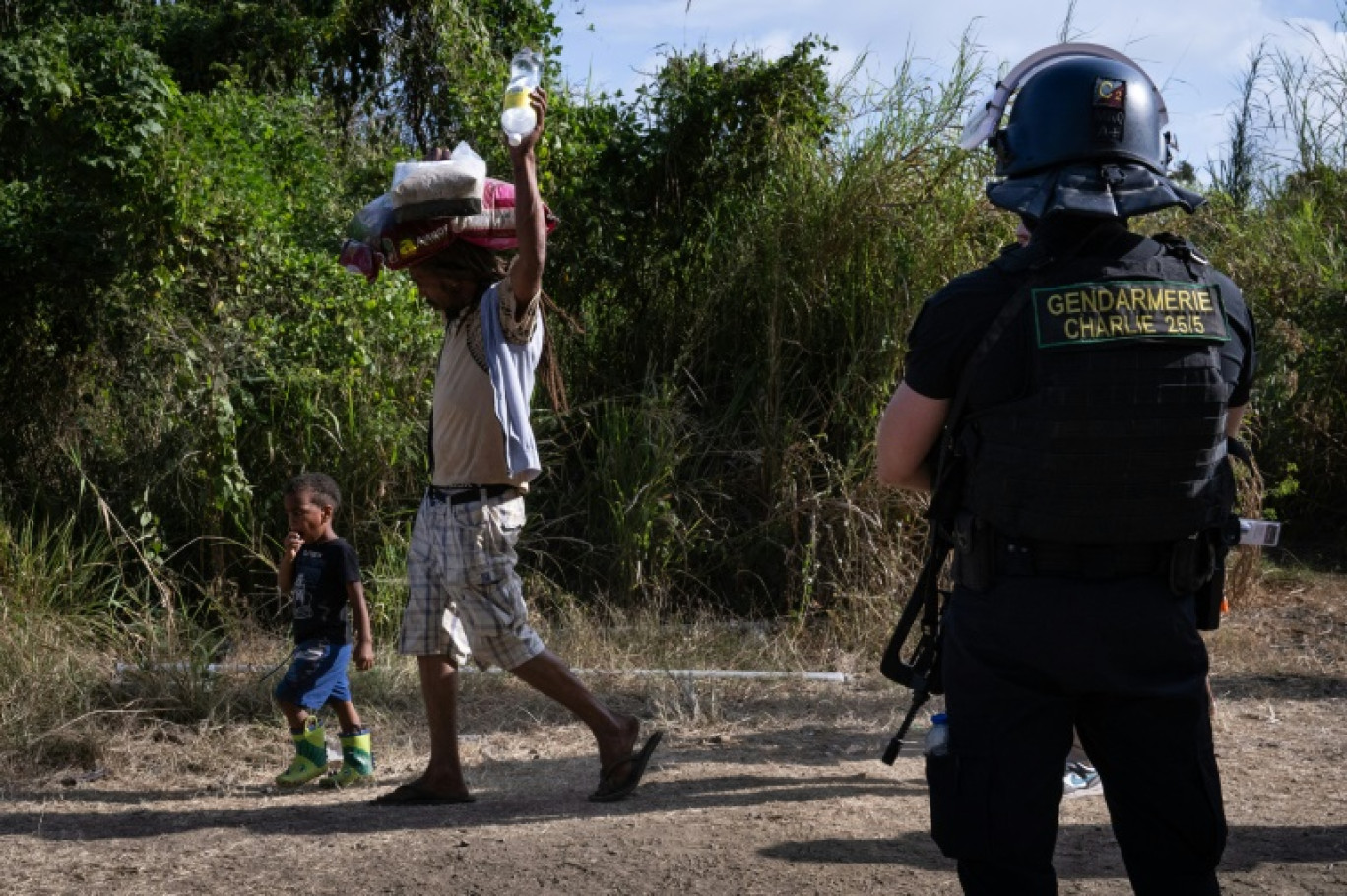 Un habitant, transportant sur sa tête des sacs de provisions, passe avec des enfants, devant un gendarme à un barrage routier à Saint-Louis, au sud de Nouméa,  le 23 septembre 2024 en Nouvelle-Calédonie © SEBASTIEN BOZON