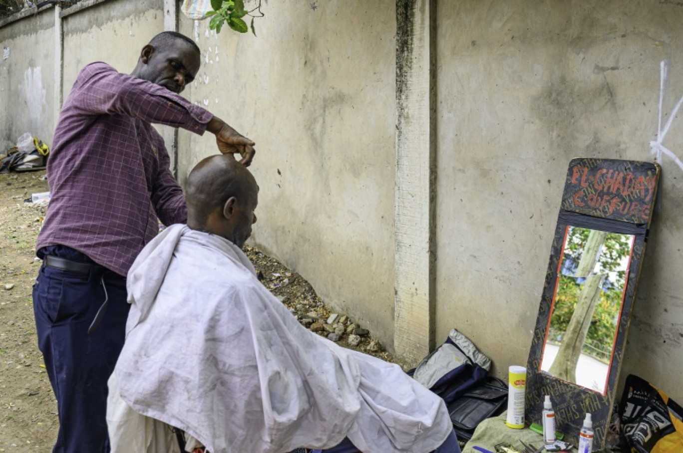 Papy Kalume, un coiffeur de rue, coupe les cheveux d'un client sur le trottoir à Kinshasa, le 17 octobre 2024 en RDC © Hardy BOPE