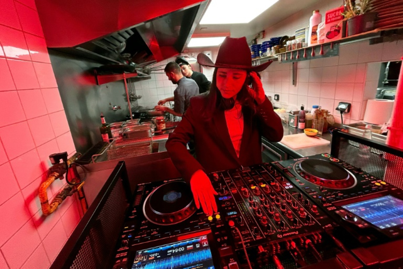 La DJ Carla Mo (c) dans les cuisine d'un restaurant lors du spectacle musical "Let Him Cook" diffusé sur les médias sociaux, pendant que le cuisinier Enzo Franceschi (g) prépare un plat sous le regard du fondateur du spectacle Théo Ferrato (d), le 9 décembre 2024 à Marseille © Viken KANTARCI